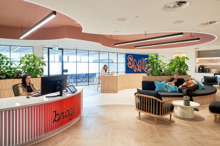 Modern office lobby with a curved reception desk labeled 'braze' where a woman is working on a computer, a casual seating area with a man using a phone, plants, and a wall sign that says SMILE in colorful letters.