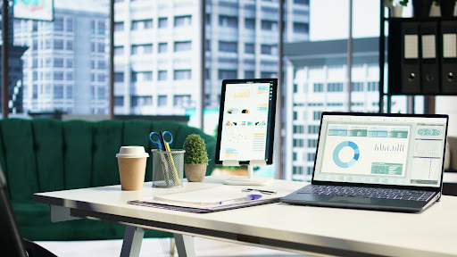 Modern office desk with laptop and tablet displaying analytics dashboards, coffee cup, notebook, and city skyline view through large windows