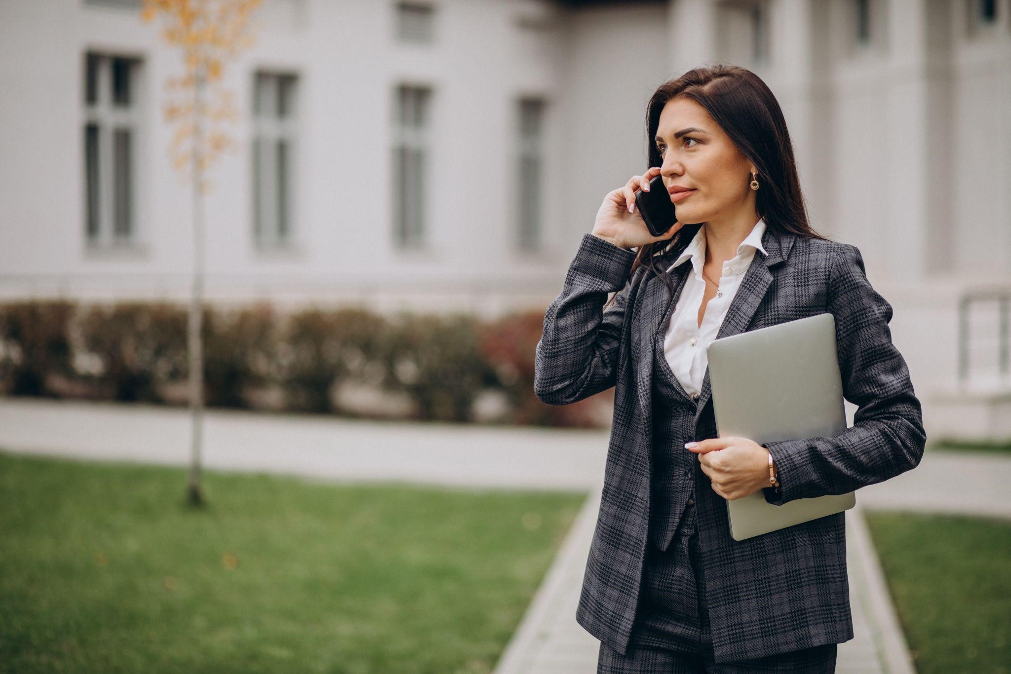 Businesswoman in suit talks on phone outdoors, holding laptop
