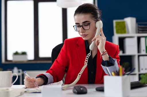 Businesswoman in red blazer taking notes while on phone call at office desk, managing tasks and communication in a professional workspace