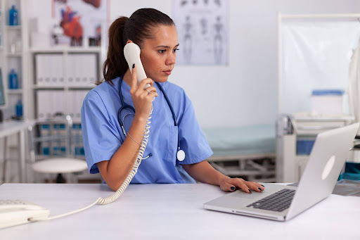 Healthcare professional in scrubs answers phone while working on laptop in clinic, managing patient communication and scheduling in medical office