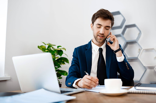 Businessman in office speaks on phone while writing notes, using laptop and managing tasks at desk with coffee and documents nearby