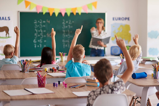 Students raising hands while teacher explains lesson at chalkboard, engaging young learners in an interactive school environment