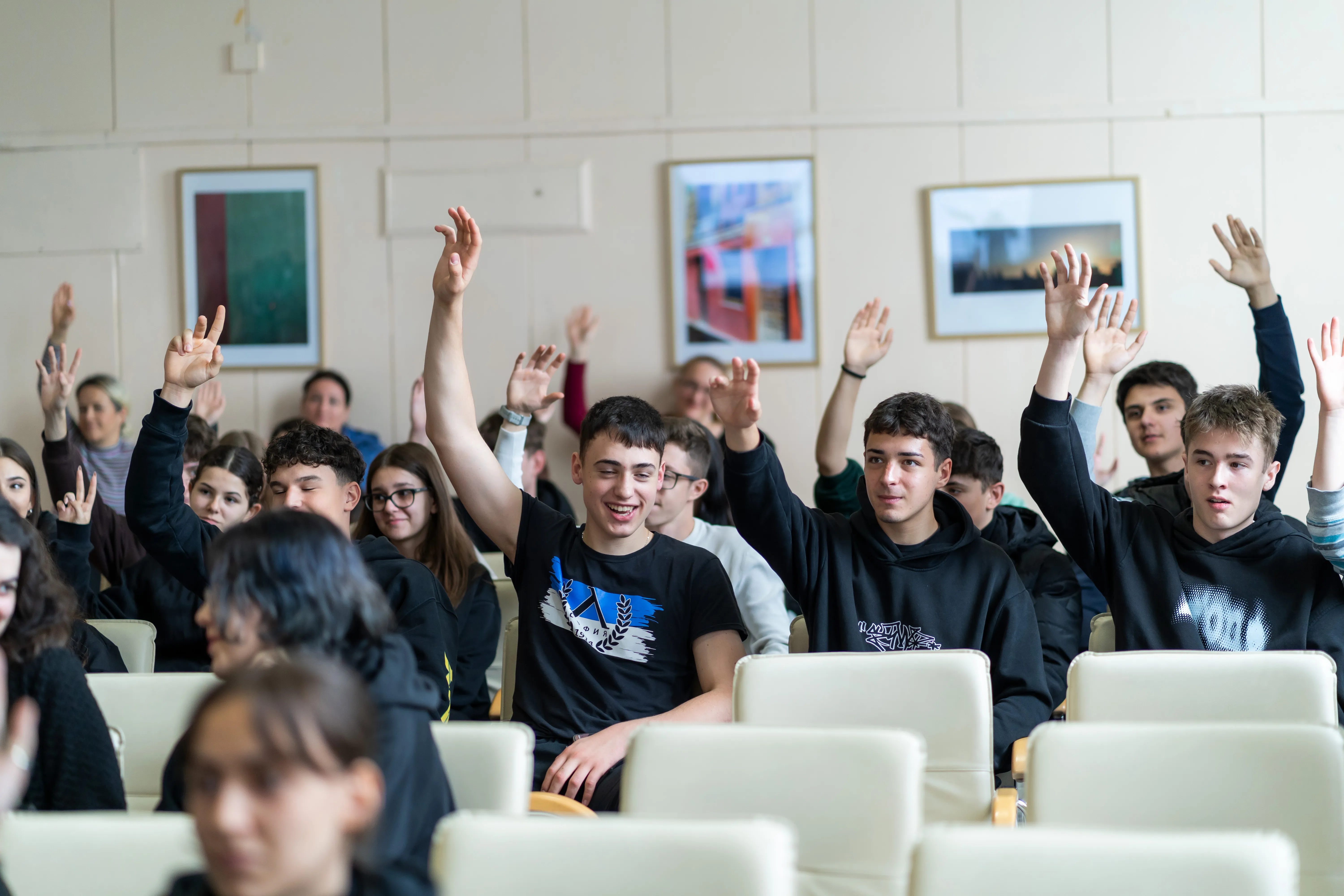Група ученици са седнали в зала за презентации. Ръцете им са вдигнати и се усмихват.