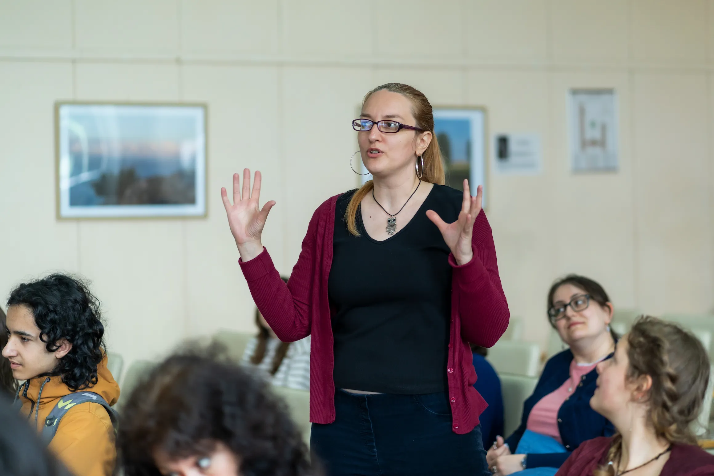 Ученичка, облечена в тъмна тениска и червена жилетка се е изправила в залата сред своите съученици и с разперени ръце обяснява нещо.