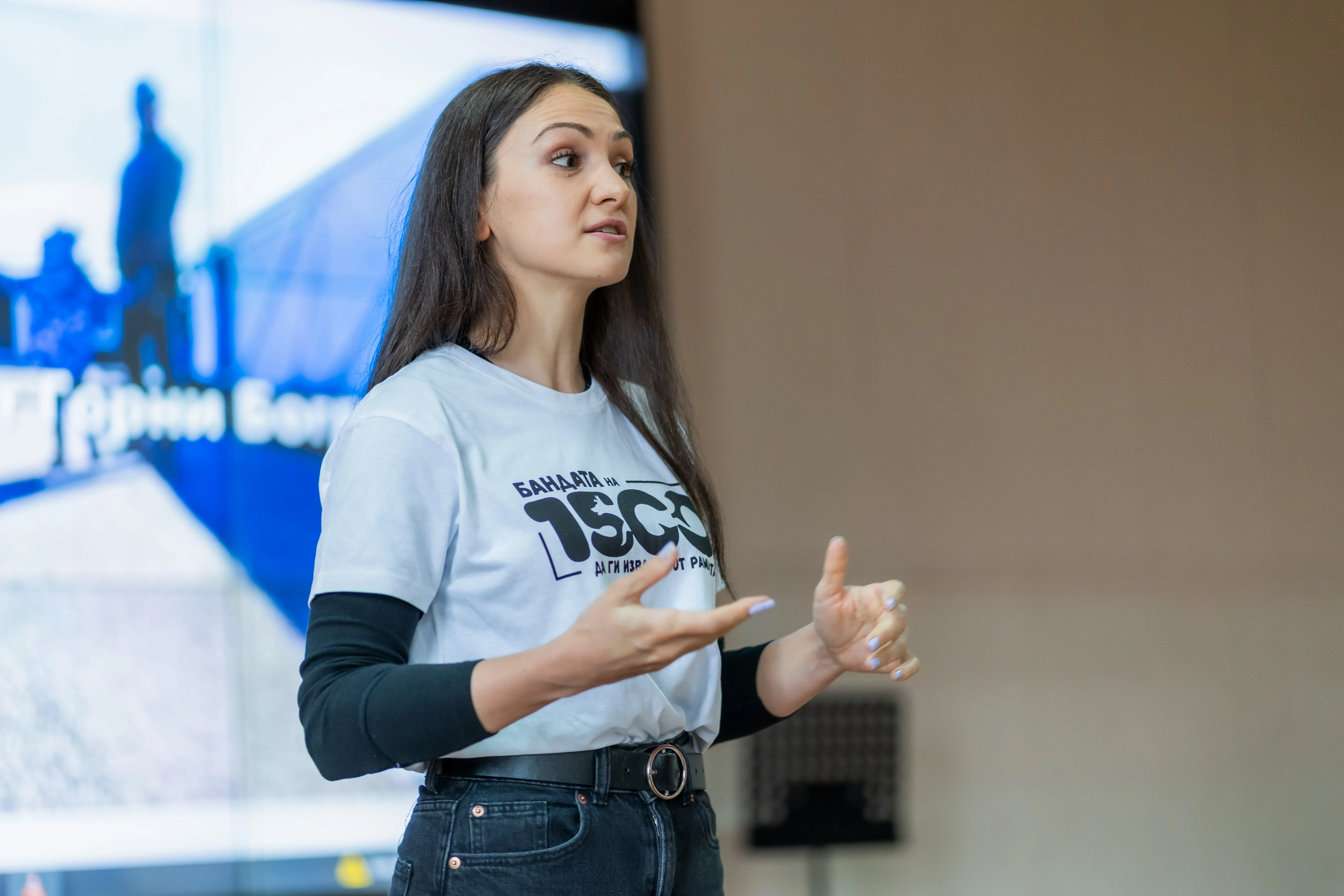 Млада доброволка на Бандата, облечена с бяла тениска с логото на организацията, обяснява разпалено каузата си. Зад нея има слайд с презентация.