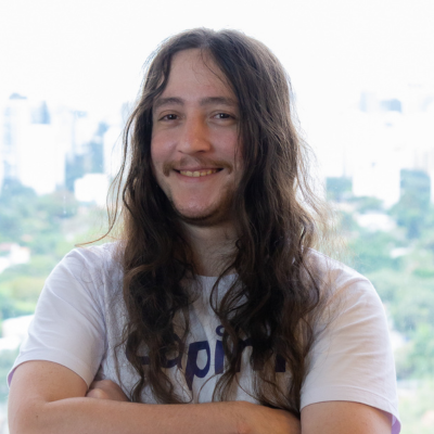 Imagem mostra Marcelo Lutz, um homem branco e alto, de cabelos castanhos, vestindo de camisa azul e segurando um microfone. Atrás dele, há um monitor exibindo a logomarca da Capim.
