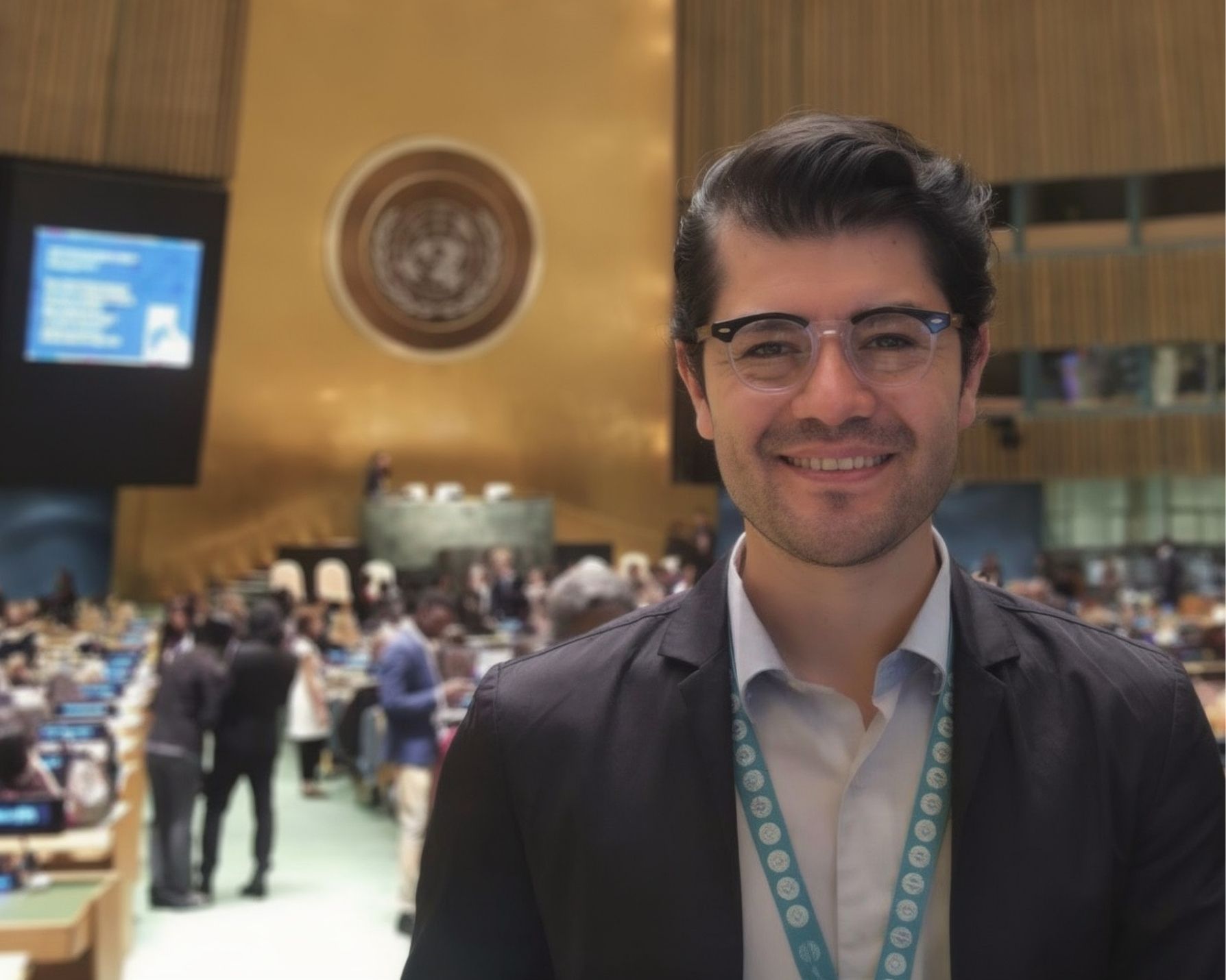 Persona con gafete en un auditorio de la ONU, de pie en primer plano con el rostro desenfocado, mientras al fondo se ve el emblema de las Naciones Unidas y varias personas reunidas en una sesión.