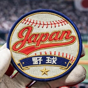 Puede incluir: Parche bordado con un diseño de béisbol. El parche presenta la palabra "Japan" en escritura roja, caracteres japoneses y una estrella dorada sobre una pancarta azul, sobre un fondo de béisbol blanco.