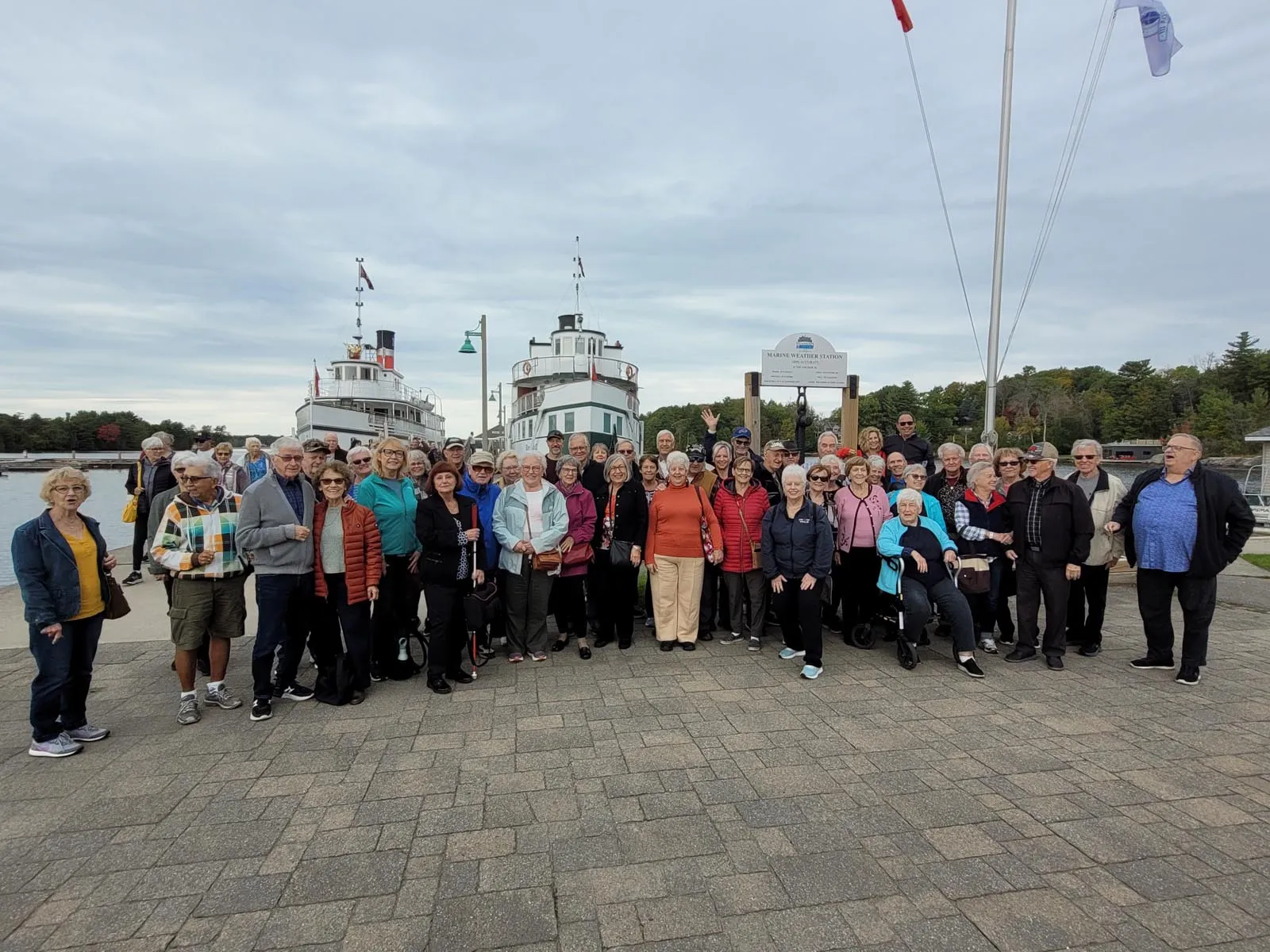 Large group of older adults posing outdoors near docked boats and a marine weather station sign.