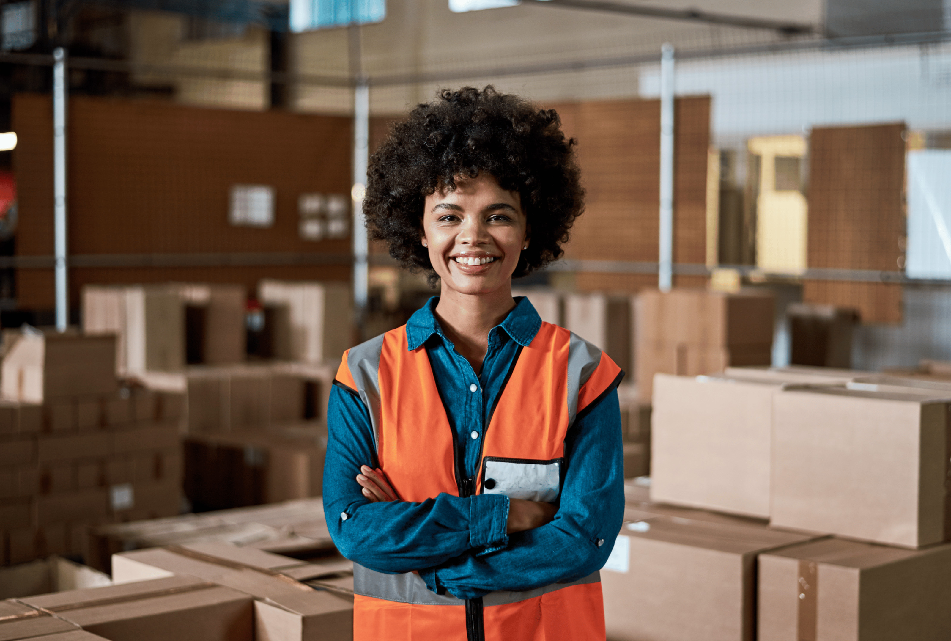 Smiling warehouse worker wearing an orange safety vest standing with arms crossed among stacked cardboard boxes.