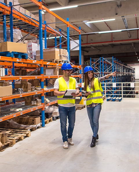 Two warehouse workers wearing blue helmets and yellow safety vests reviewing inventory with a tablet inside a large storage facility.