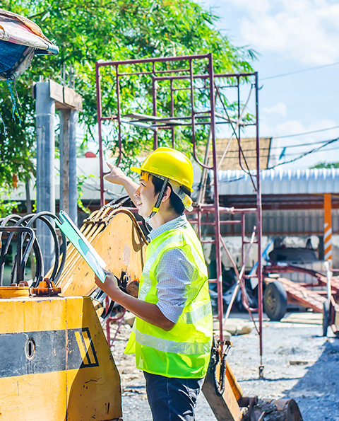 Construction worker wearing a yellow hard hat and safety vest pointing while holding a clipboard at a construction site with machinery and scaffolding.