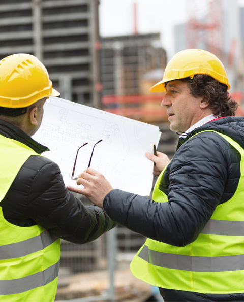 Two construction workers in yellow helmets and reflective vests reviewing a blueprint on a construction site.