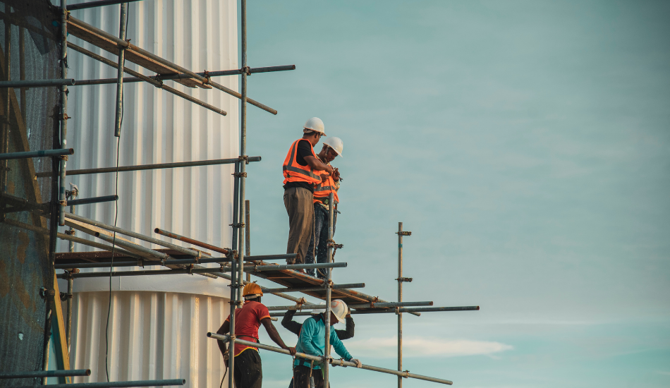 Four construction workers wearing helmets and safety vests assembling scaffolding against a large white structure under a clear sky.