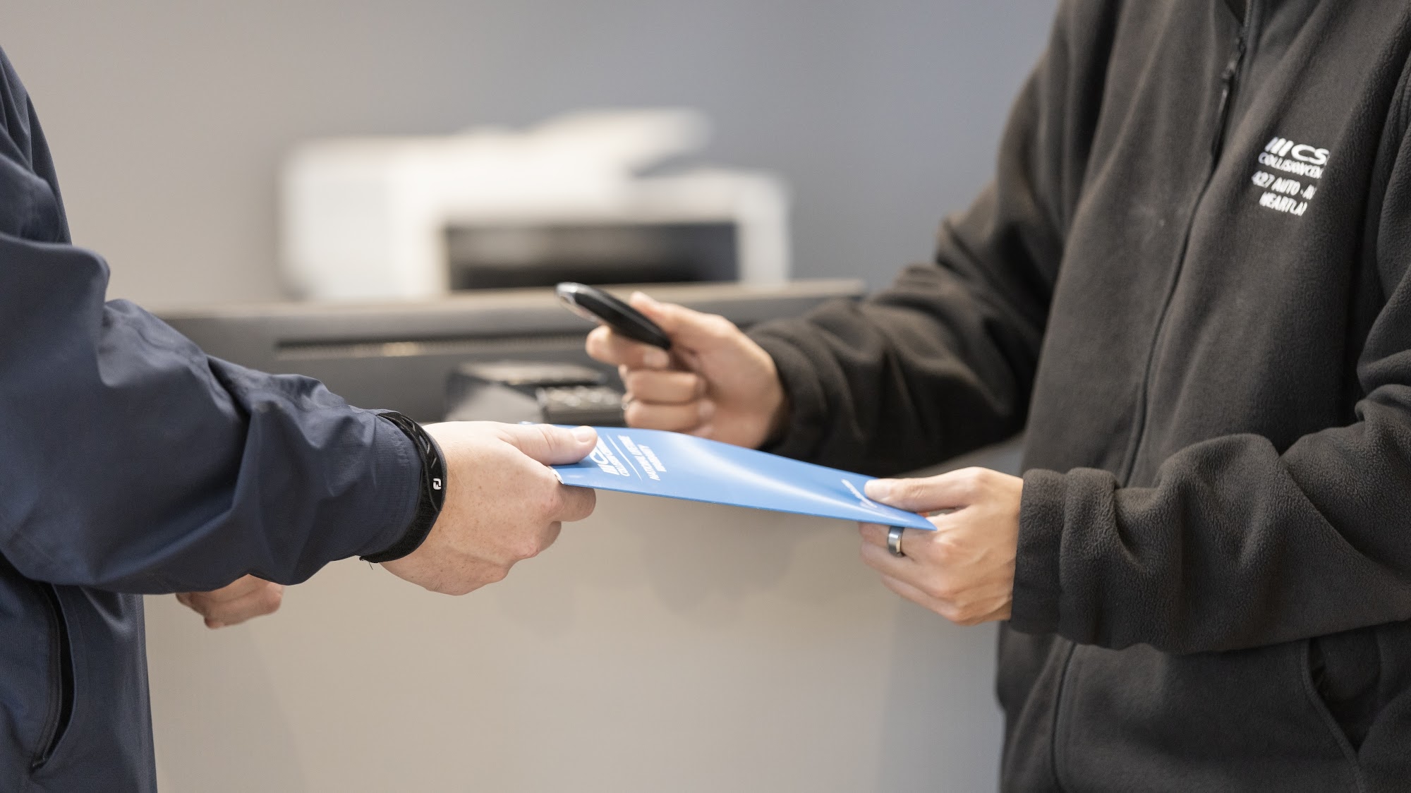 A CSN 427 Auto staff member hands a customer a blue repair folder and car key at the service counter