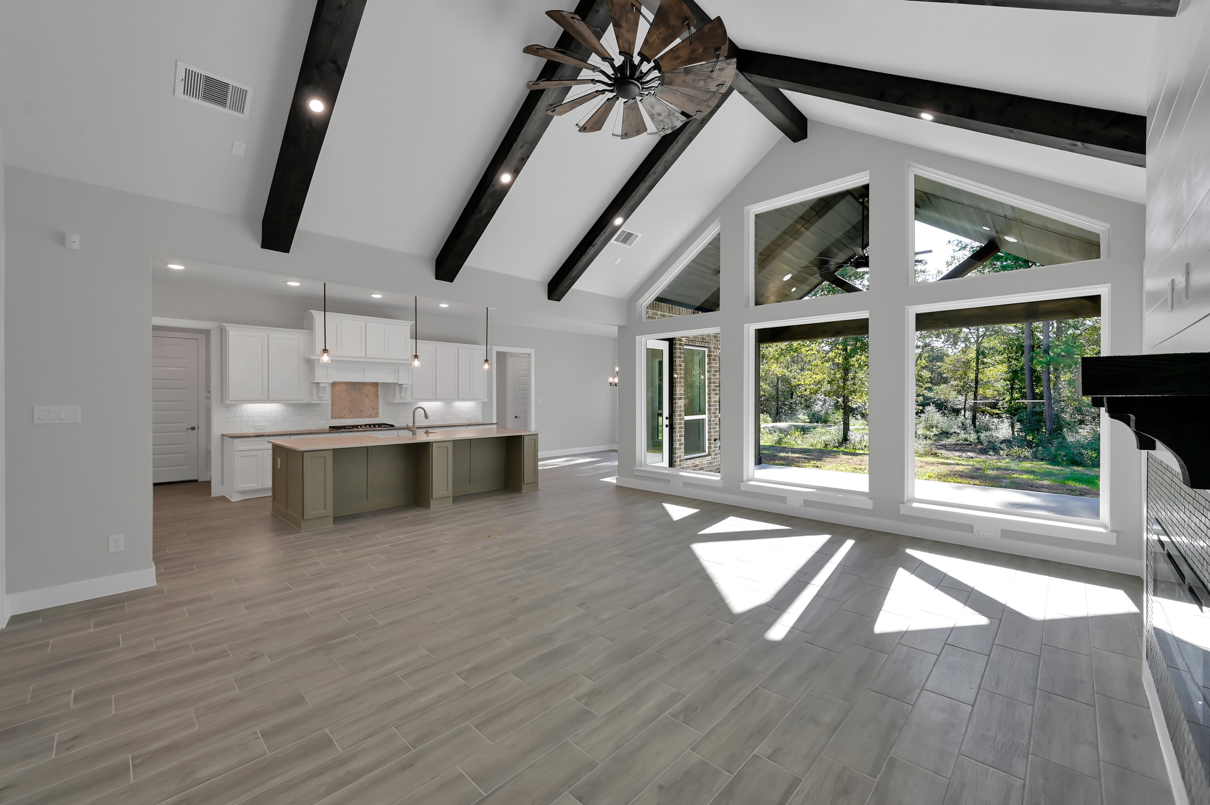 Open-concept modern farmhouse great room with a vaulted ceiling, dark exposed beams, a windmill-style ceiling fan, and large windows over a white-and-sage kitchen
