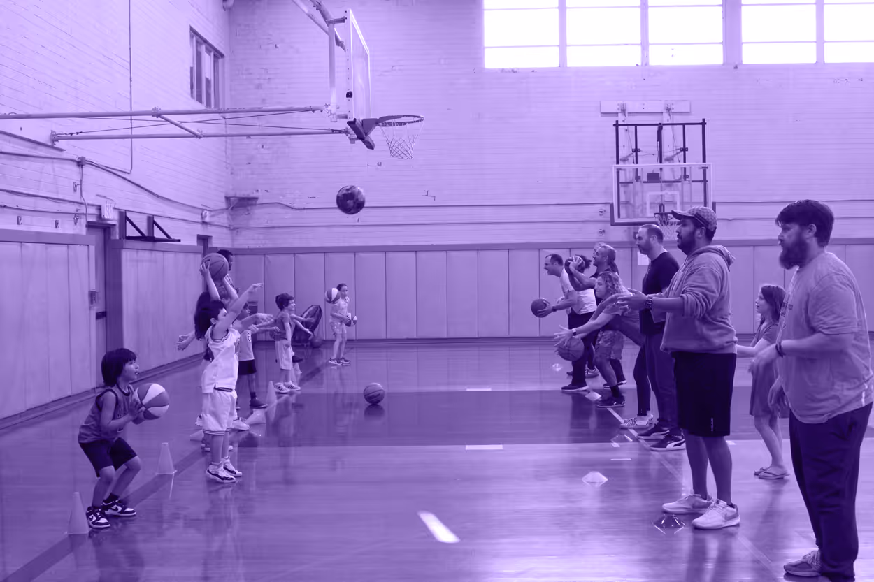 A group of children and adults participating in a Youth Basketball Clinic, practicing shooting baskets in a gym at the Jewish Community Center, Mid City LA. Several people are lined up, with basketballs in hand.