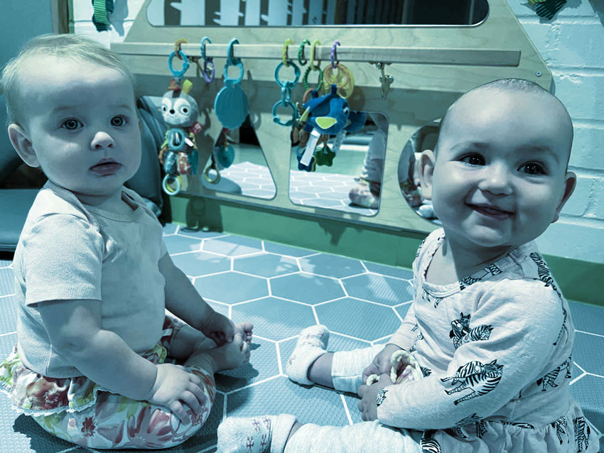 Two babies sitting on a blue play mat with hanging toys and mirrors behind them.