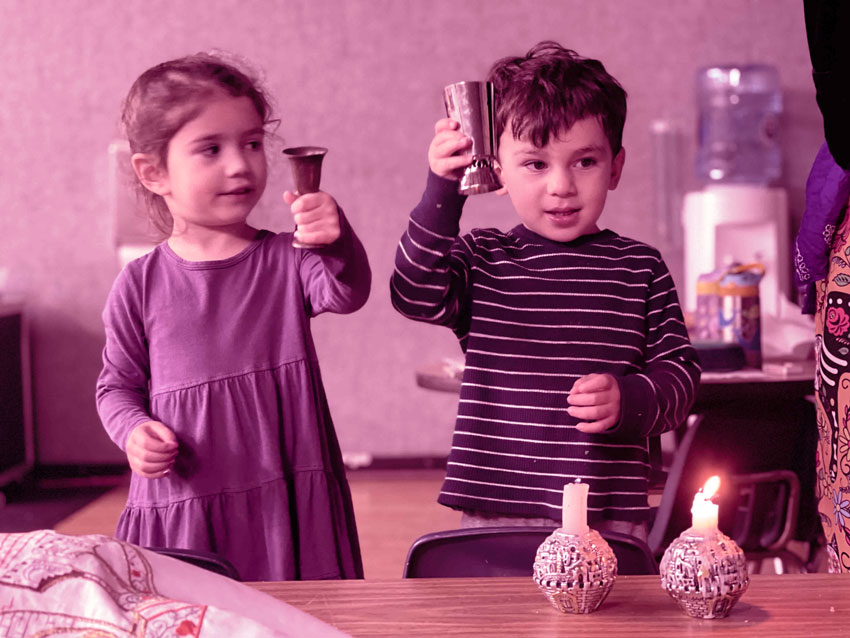 Two children holding bells, standing behind a table with two lit candles.