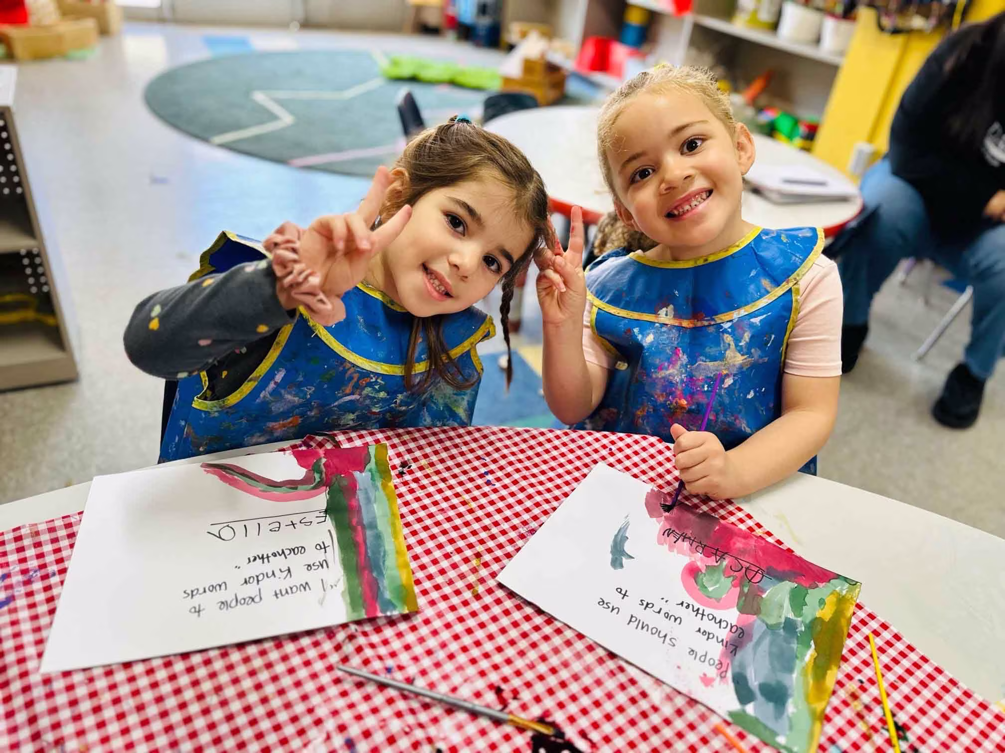 Two young children wearing blue paint aprons smiling and making peace signs while sitting at a table with painted papers.