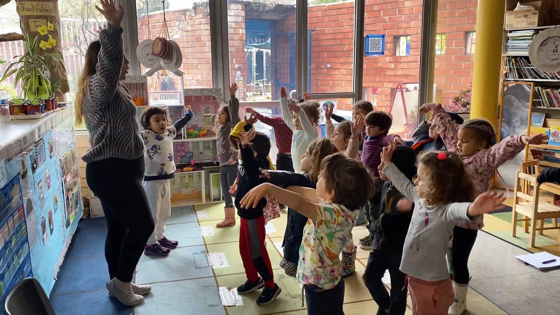Teacher leading a group of young children in a classroom activity with their arms raised.