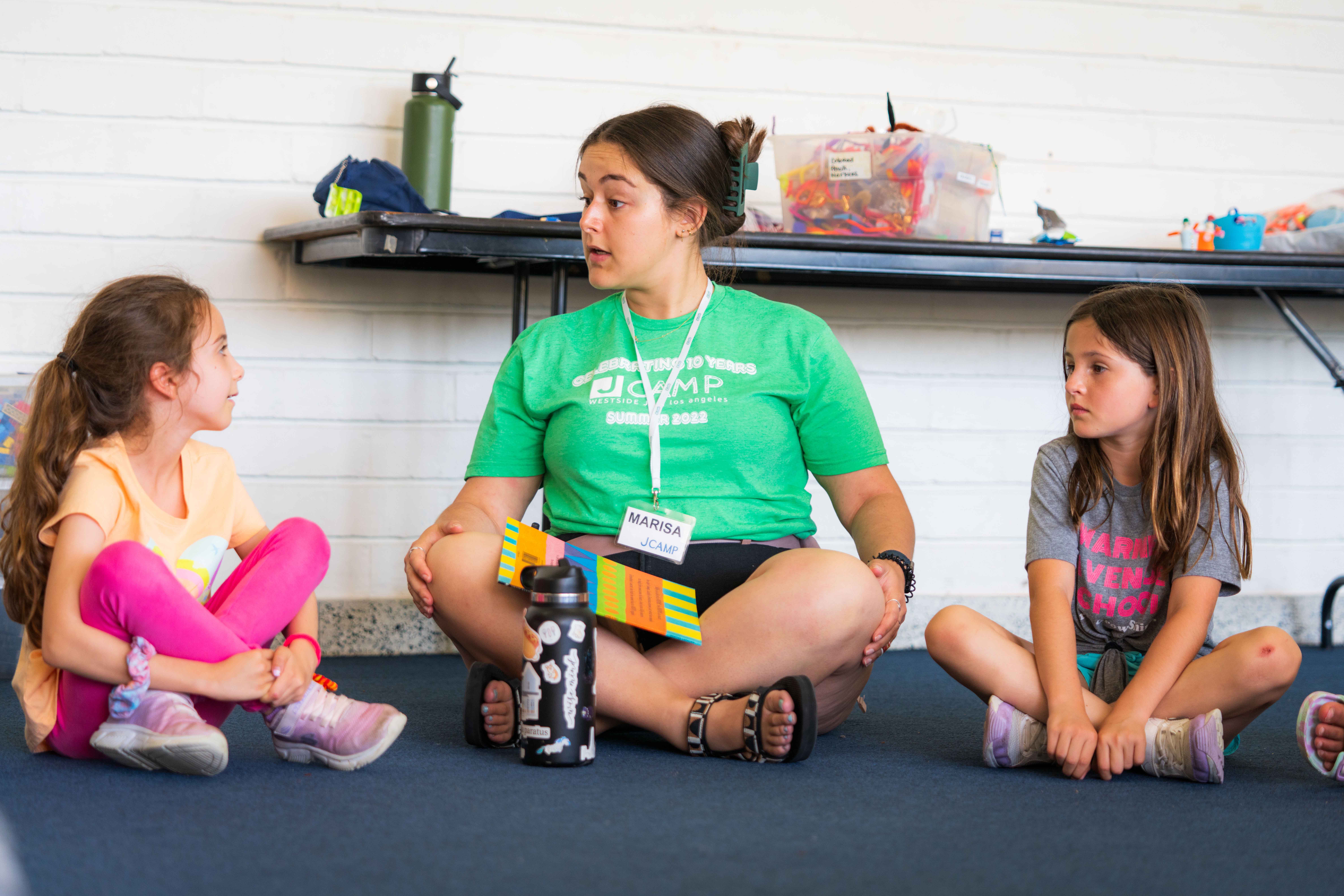 Camp counselor in green shirt sitting cross-legged on carpet floor, talking to two young girls sitting on either side.