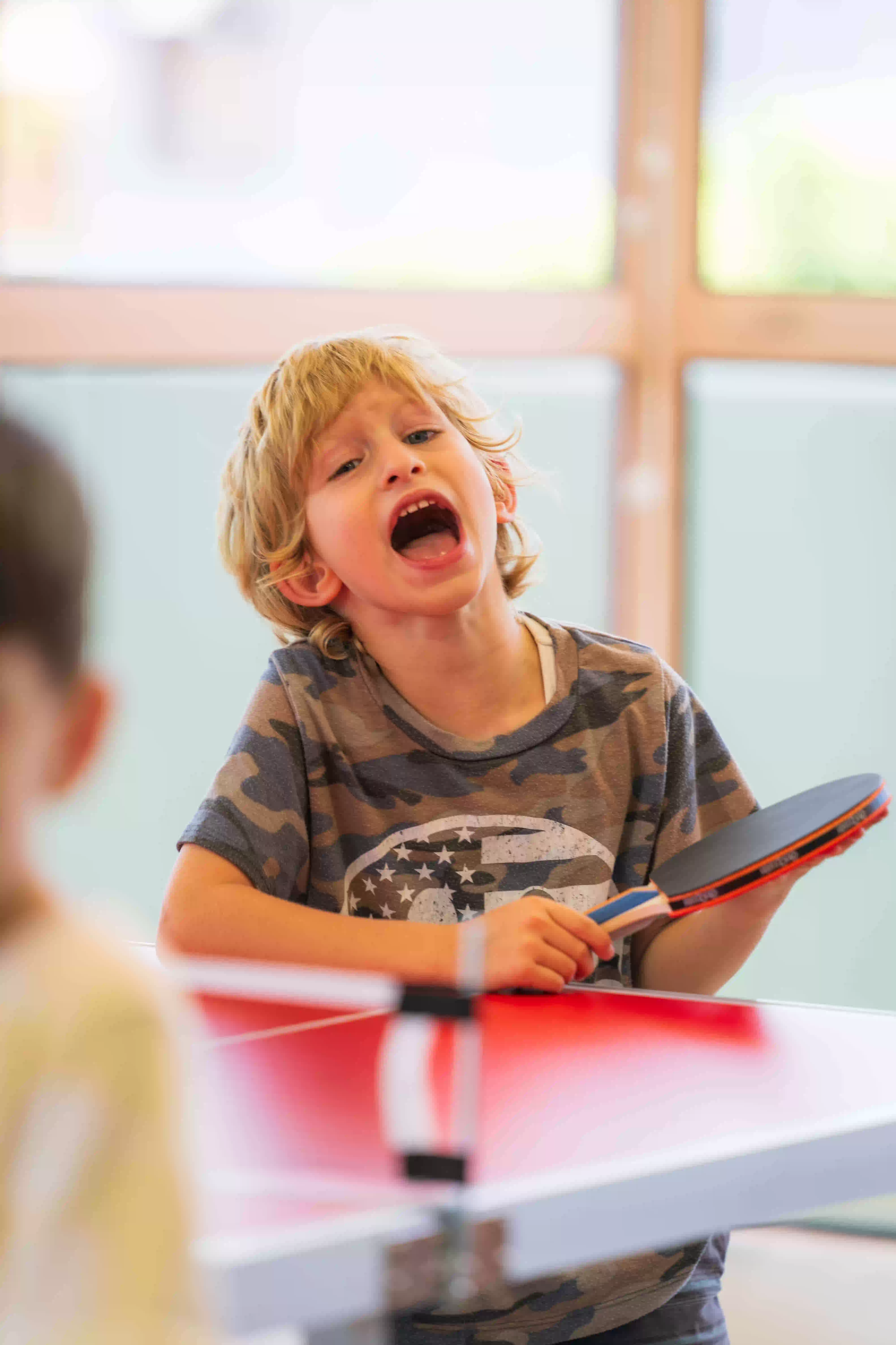 Young boy with blond hair holding a ping pong paddle and shouting during a game.