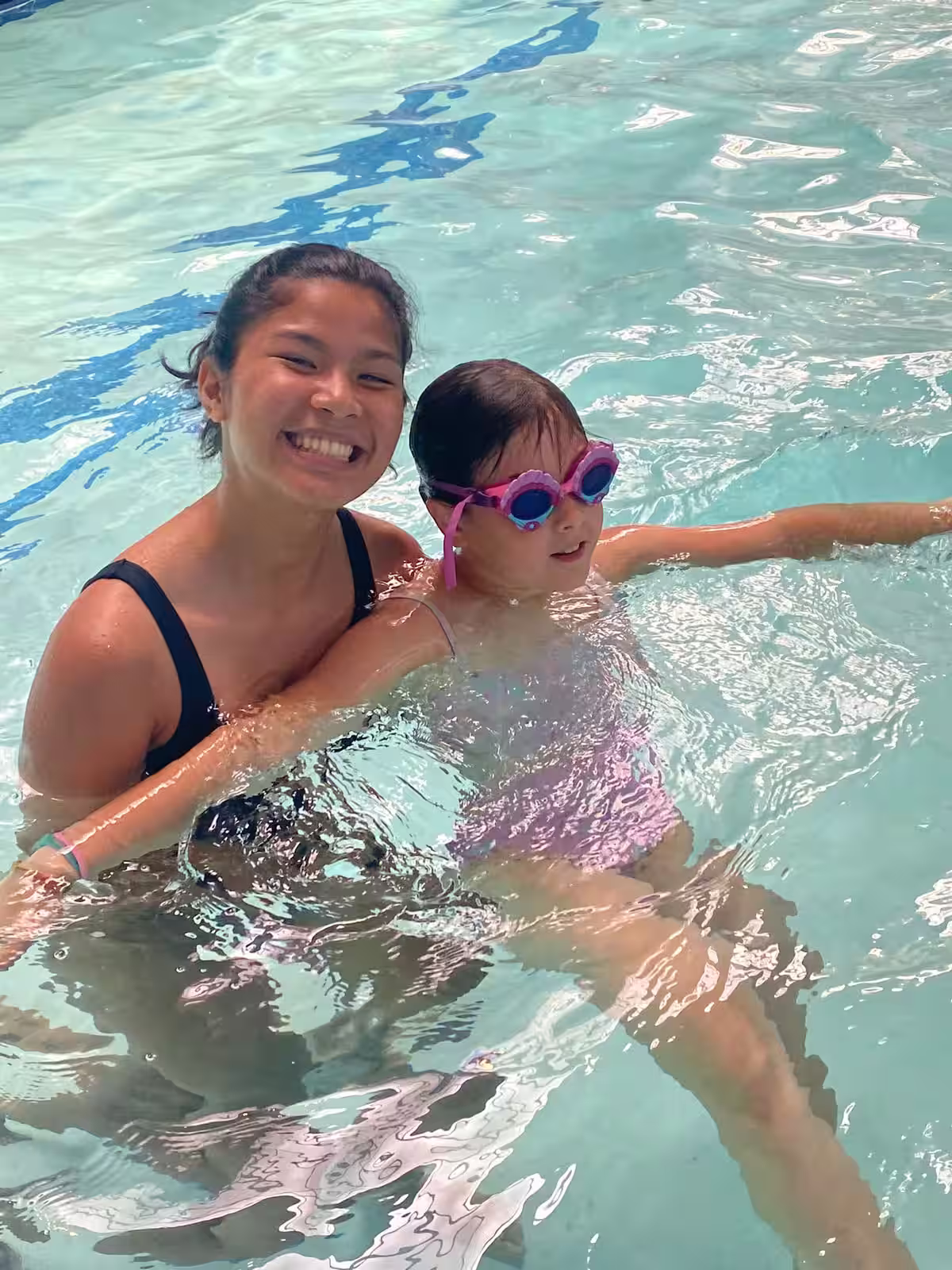 Smiling woman in a black swimsuit supports a young girl wearing pink goggles and a pink swimsuit as she floats in a pool.