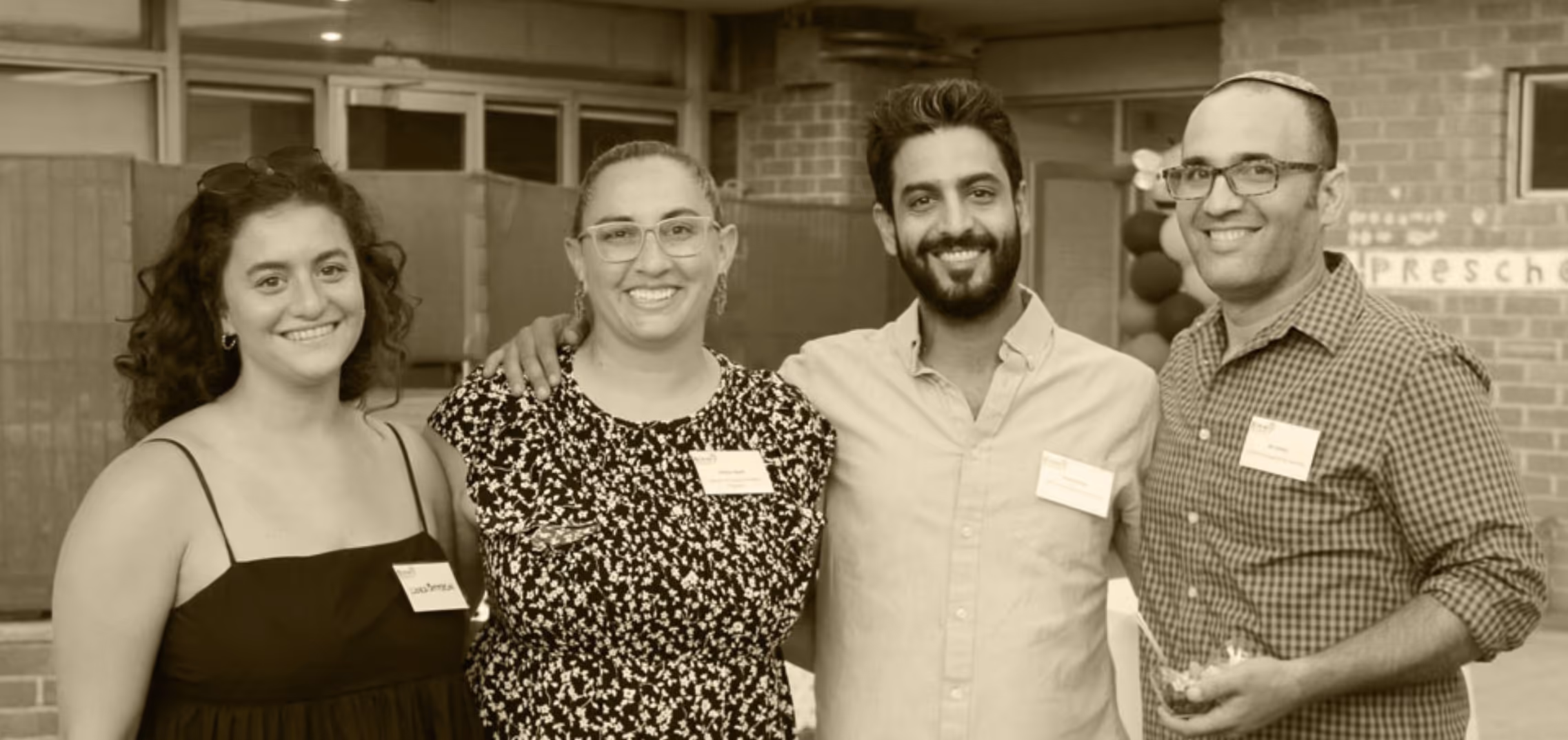Four adults smiling with arms around each other at an indoor event with name tags and balloons in the background.