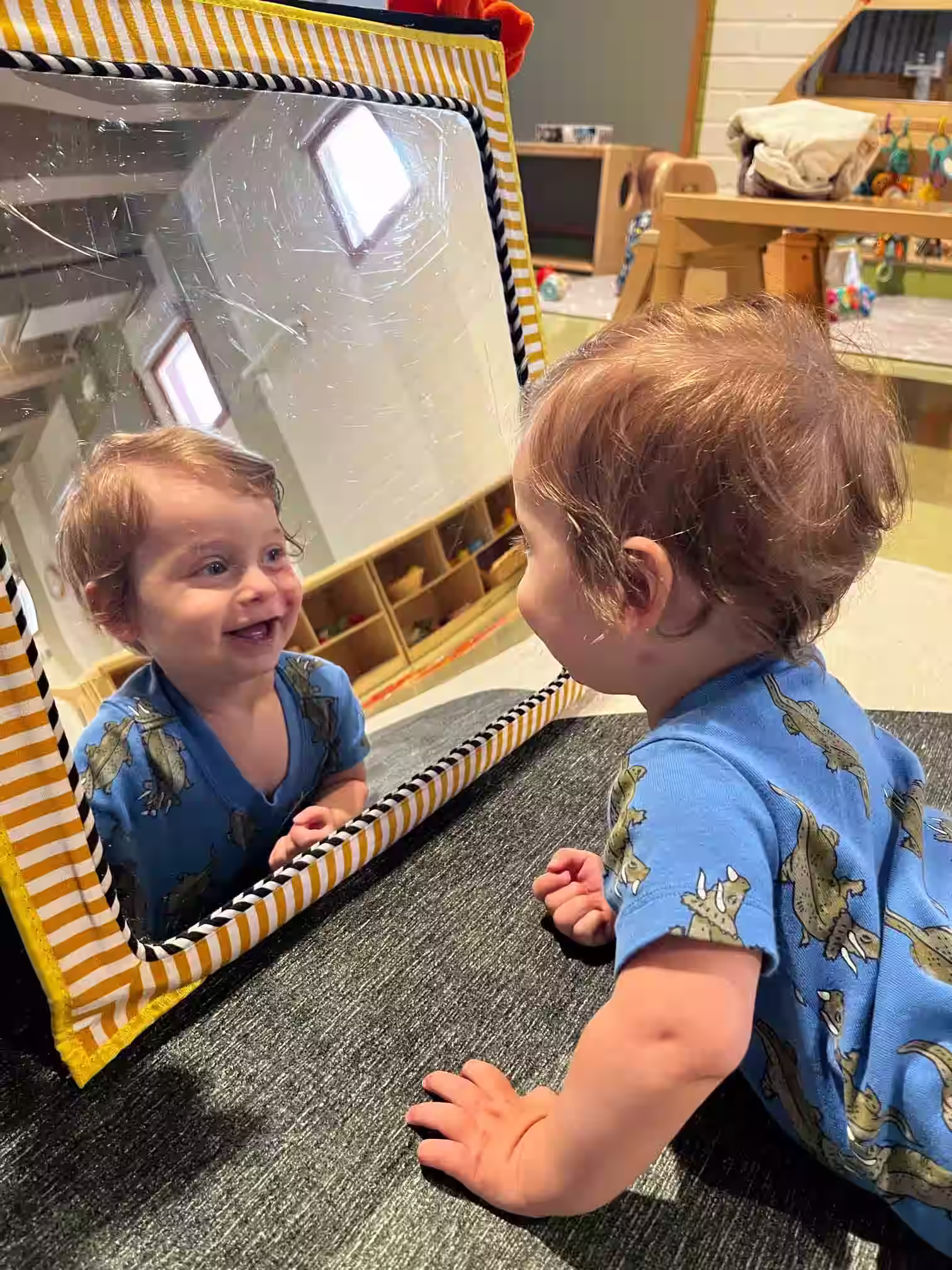 A toddler smiling and looking at their reflection in a framed mirror on the floor.