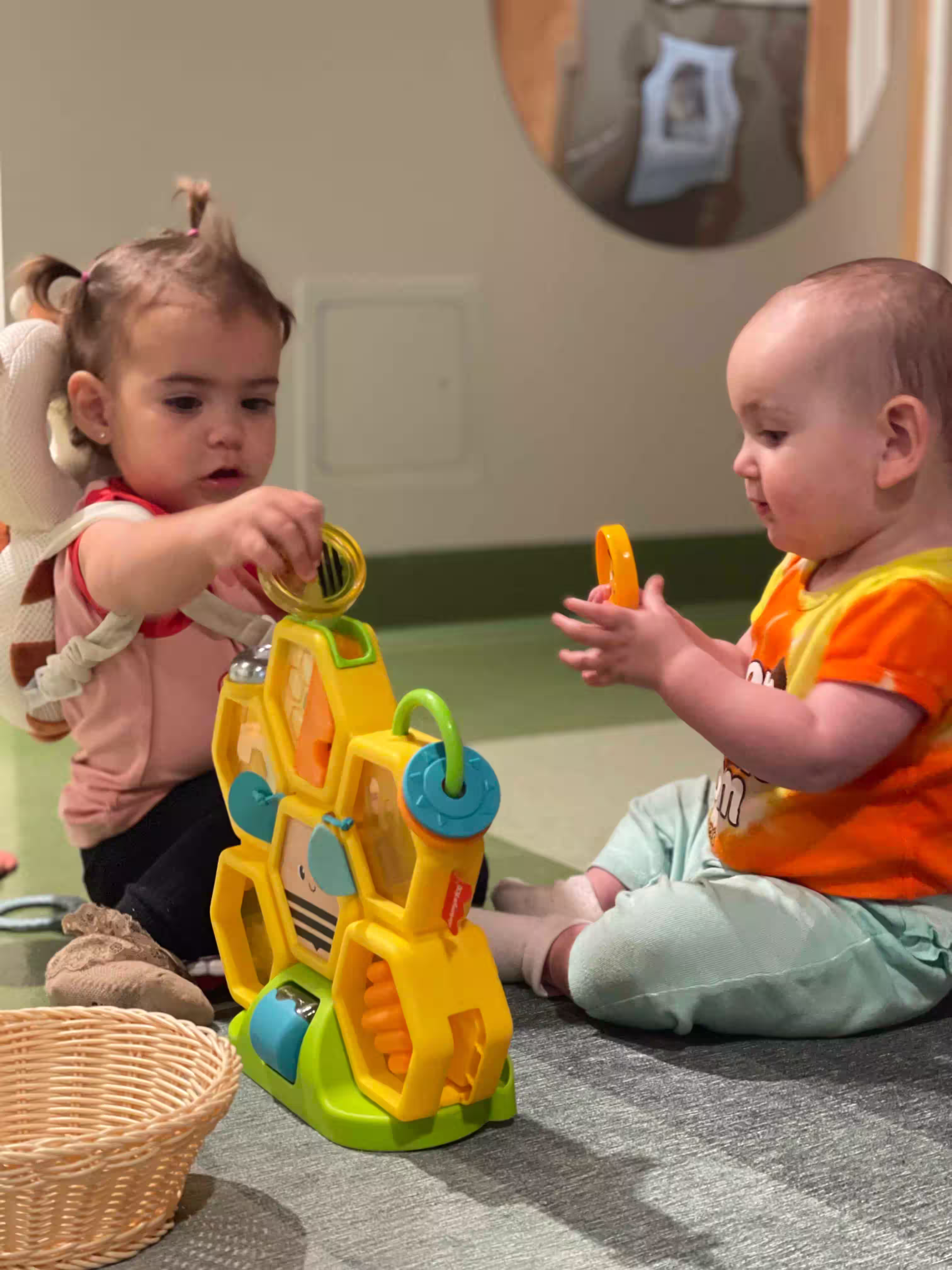 Two toddlers seated on a play mat, interacting with a yellow and green bee-shaped activity toy.