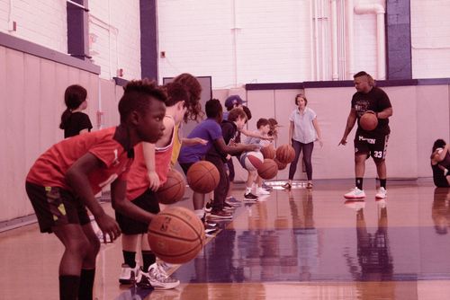 Children participating in a basketball training session at the Jewish Community Center in Mid City LA. They are lined up, dribbling basketballs, with a coach demonstrating techniques.