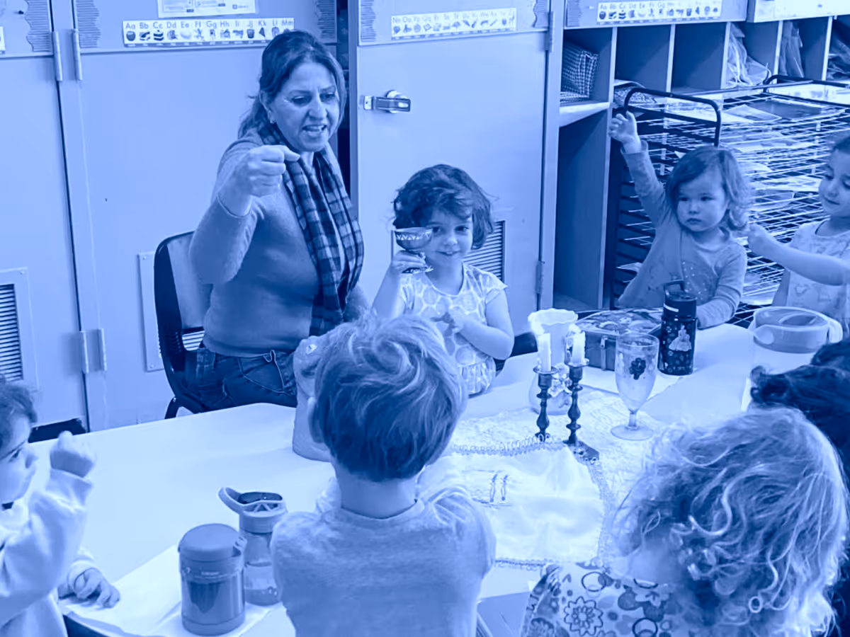 Teacher and young children sitting around a table raising their hands during a classroom activity, with two lit candles on the table.