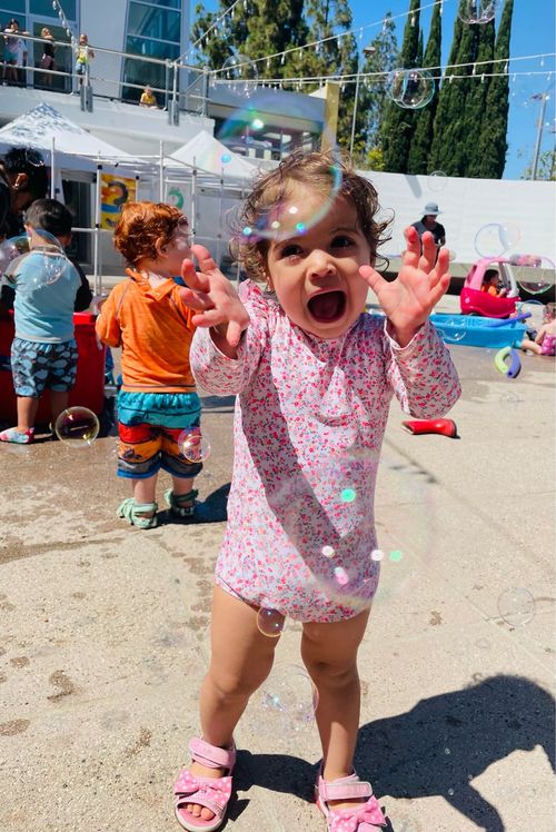 Toddler in pink floral swimsuit and pink sandals joyfully reaching out to catch bubbles outdoors on a sunny day.