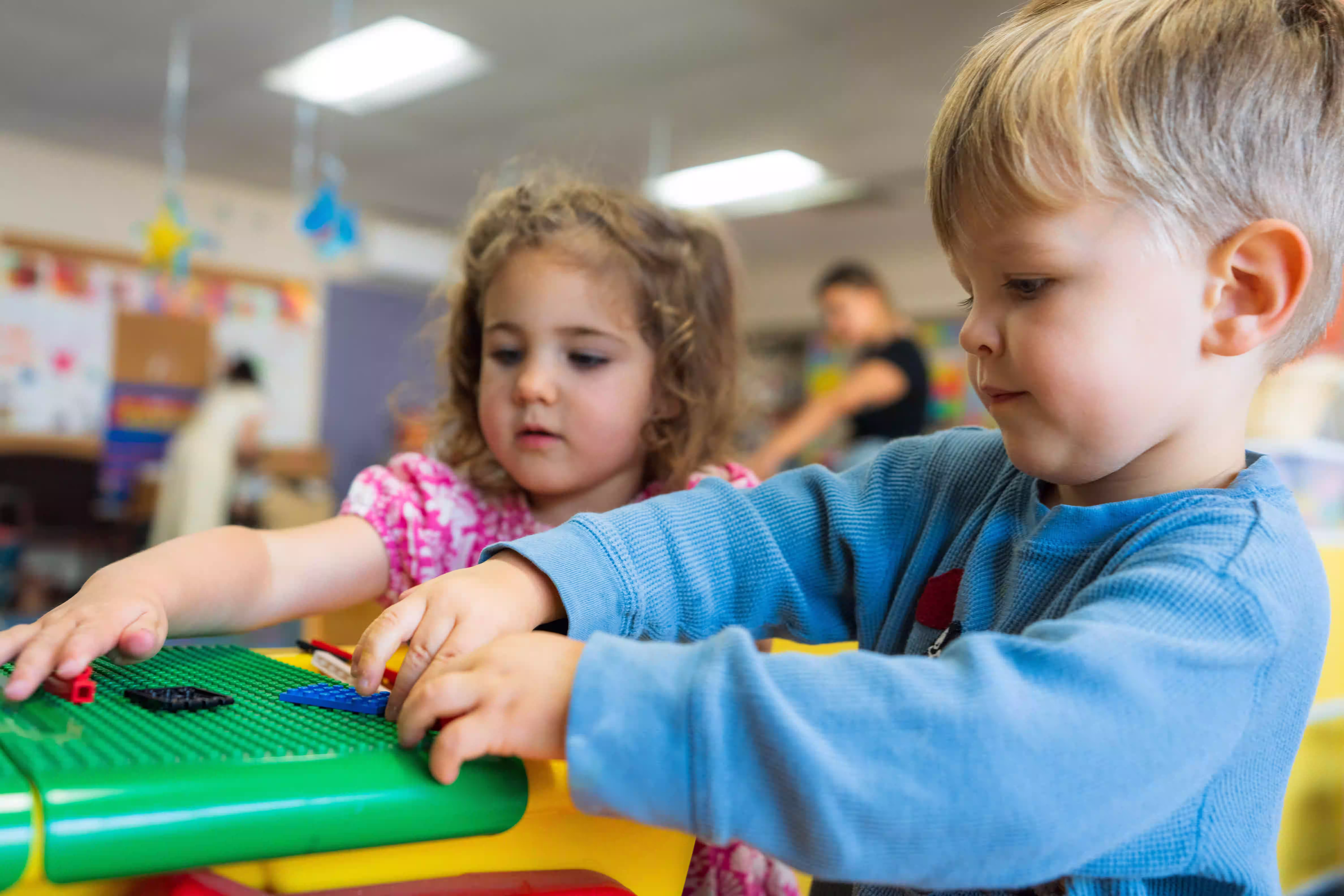 Two children playing with building blocks on a green Lego base in a classroom setting.