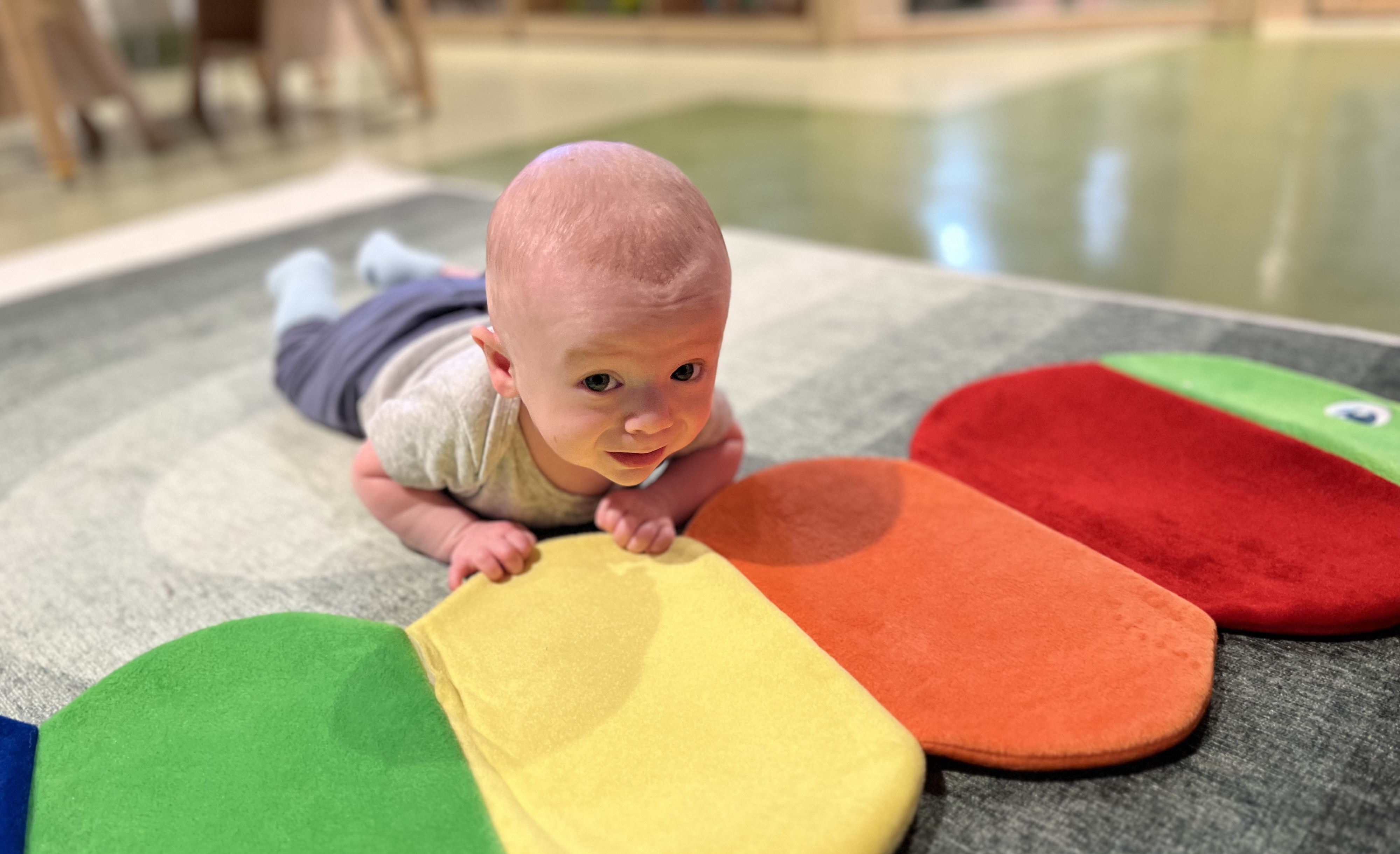 Baby lying on stomach on a gray circular rug, playing with a colorful caterpillar-shaped cushion.