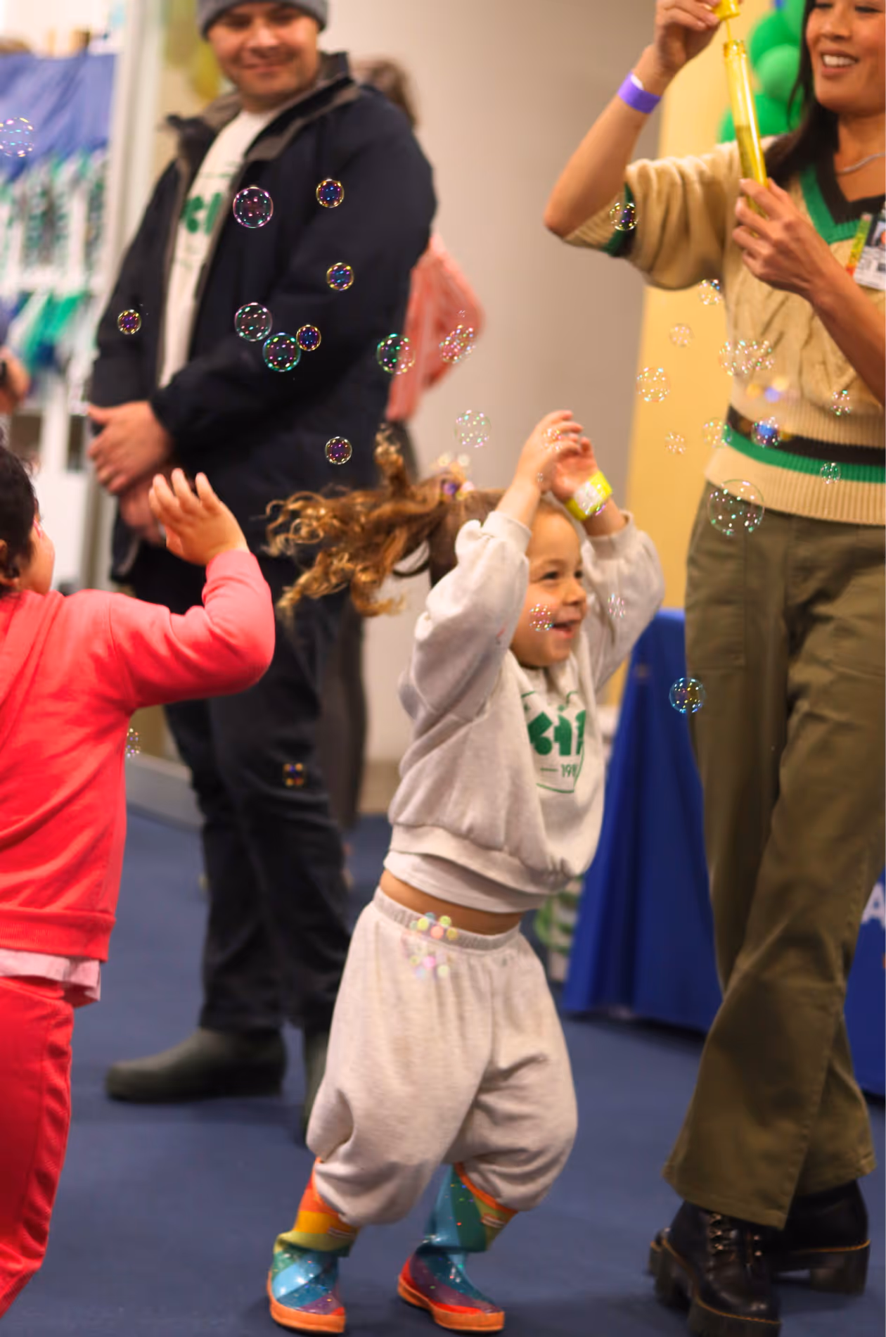 Children joyfully playing with bubbles indoors while an adult blows bubbles and a man watches smiling.