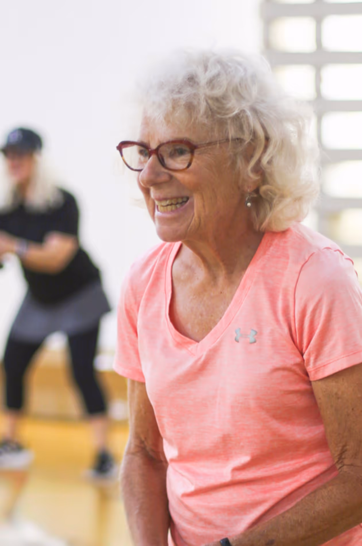 Smiling elderly woman with curly white hair and red glasses wearing a pink Under Armour shirt in a gym setting.