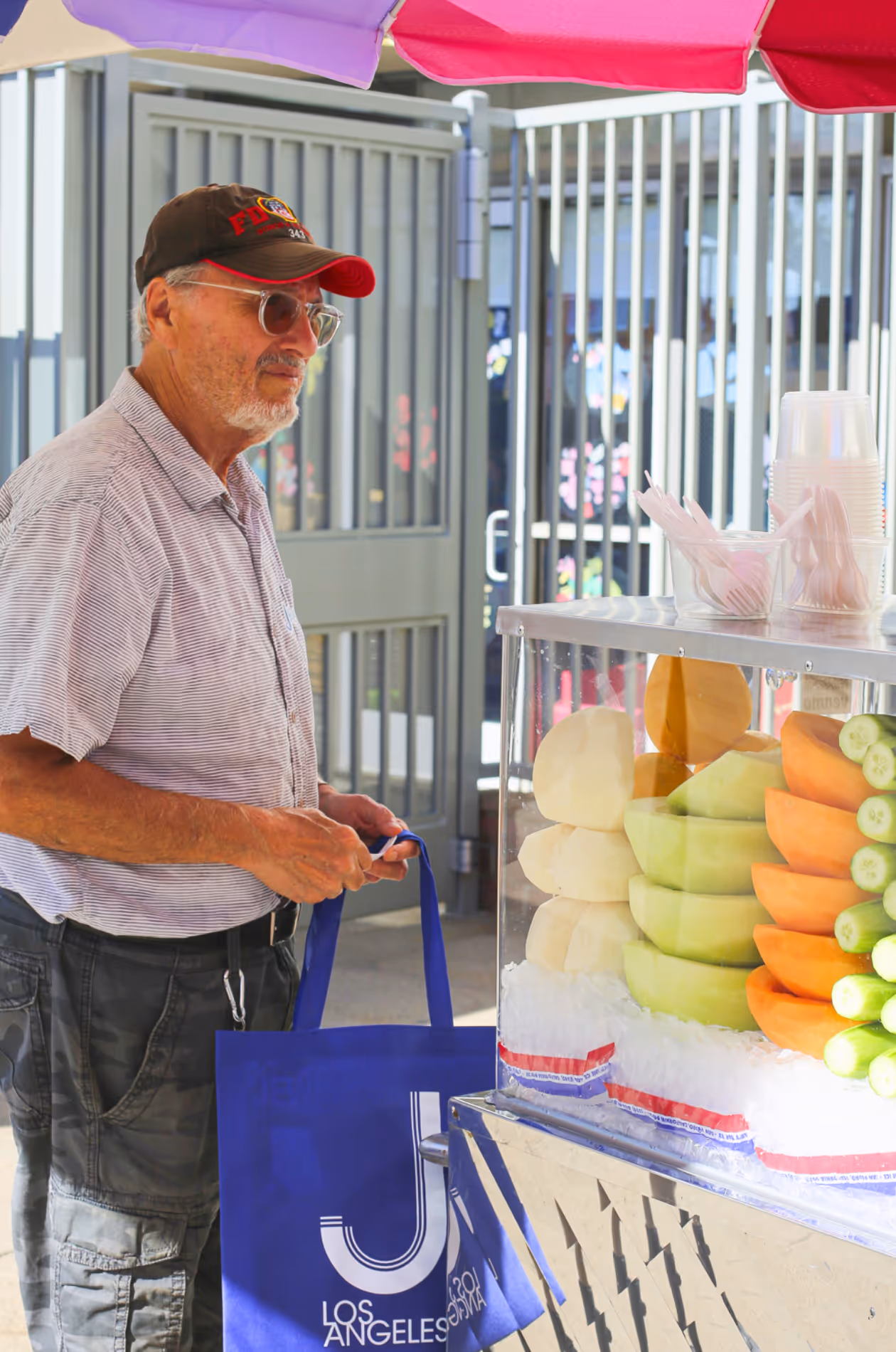 Elderly man with glasses and cap holding a blue reusable Los Angeles tote bag, standing near a fruit stand under a colorful umbrella.