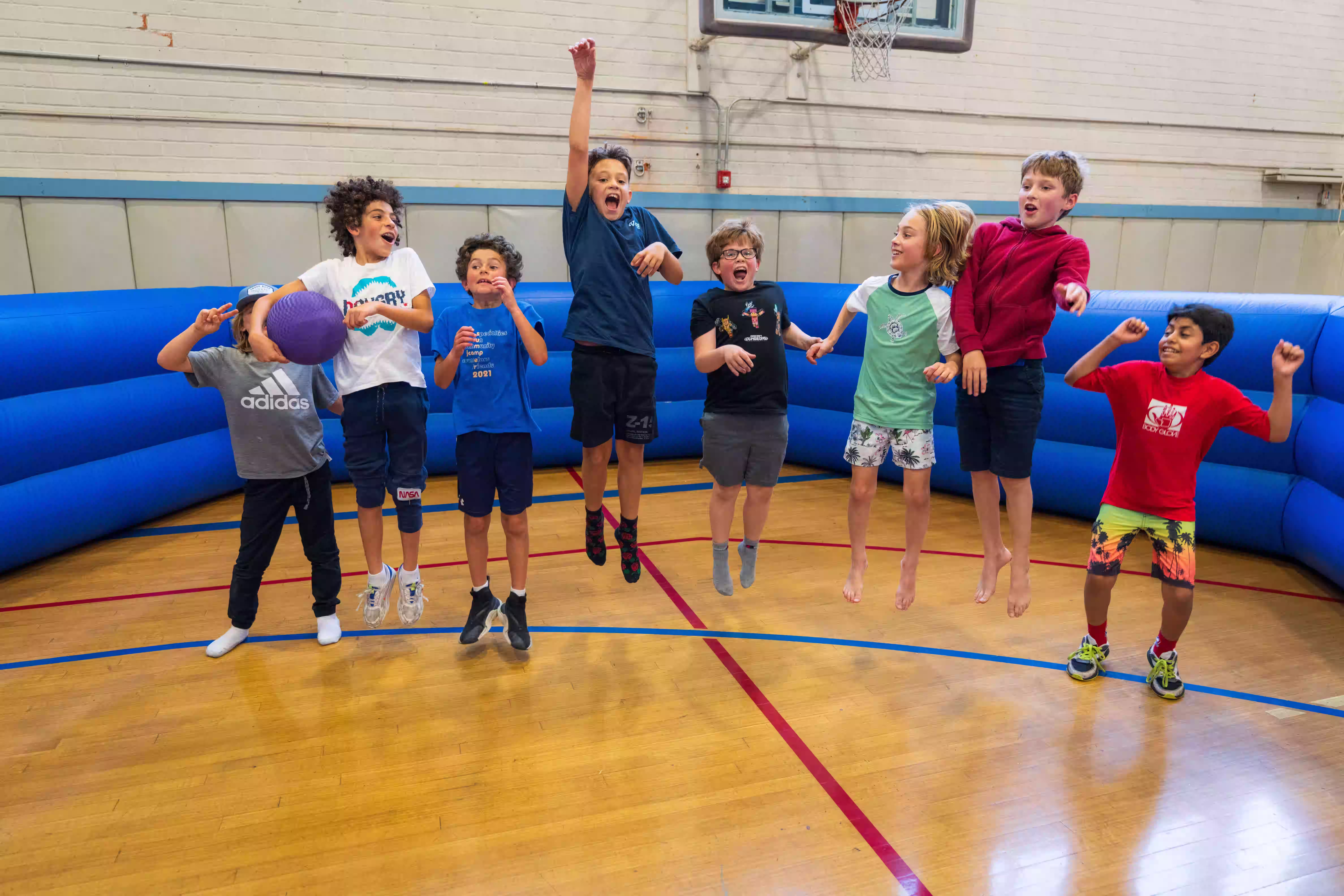 Eight boys jumping and playing inside a gym with a blue inflatable barrier and a basketball hoop.
