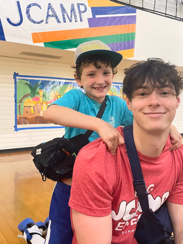A smiling young boy wearing a blue shirt and cap hugs a young man with a red JCamp shirt and sling bag inside a gymnasium.