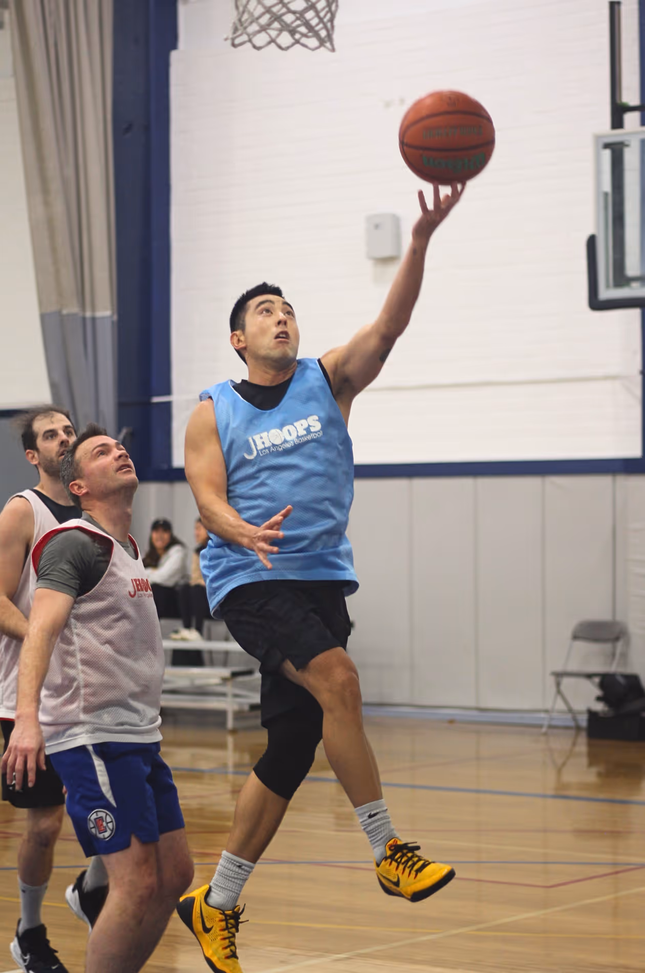 A basketball player in a blue JHoops jersey jumps for a layup while two defenders watch in an indoor gym.