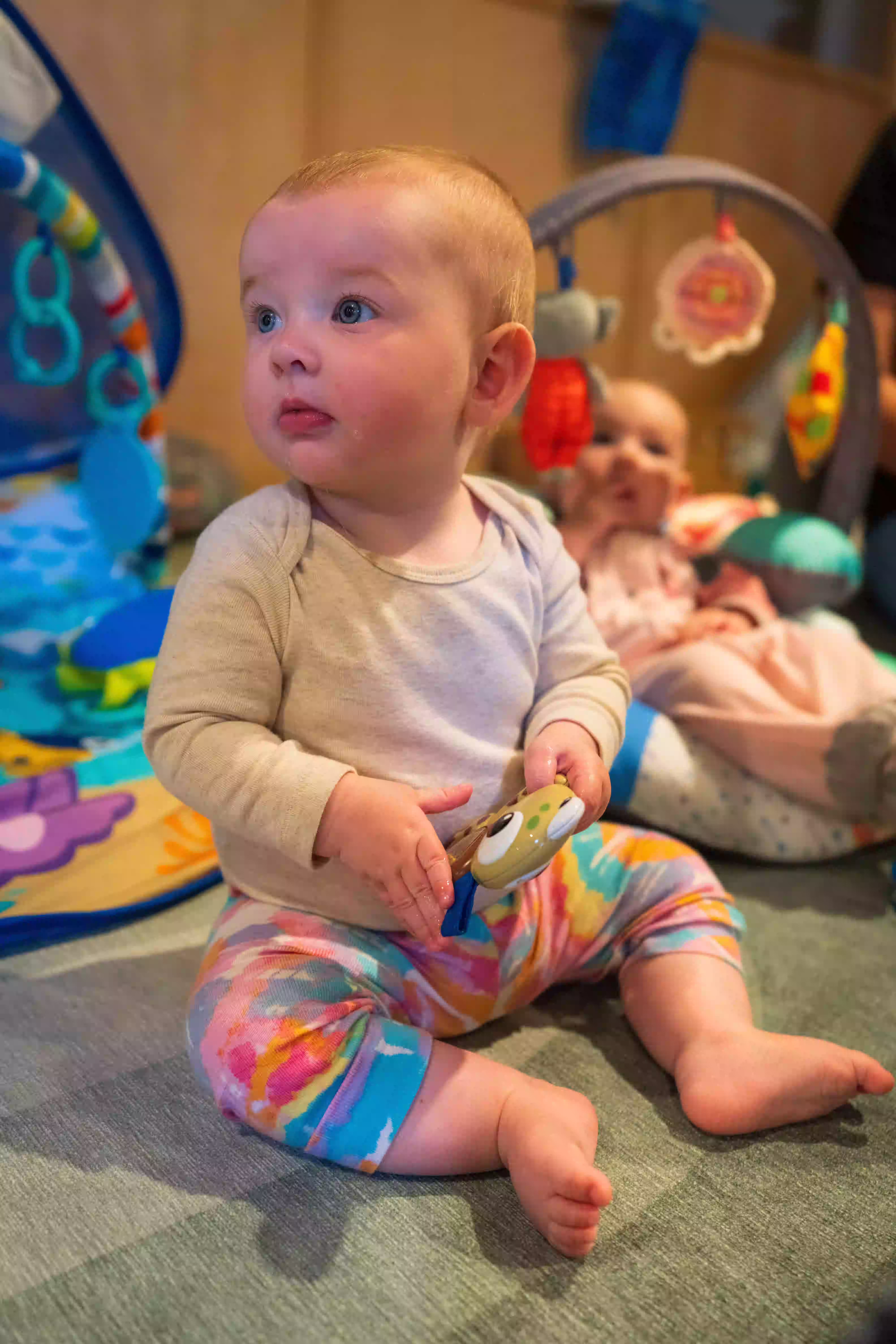 Baby in colorful pants sitting on the floor holding a toy, with another baby and play gym in the background.