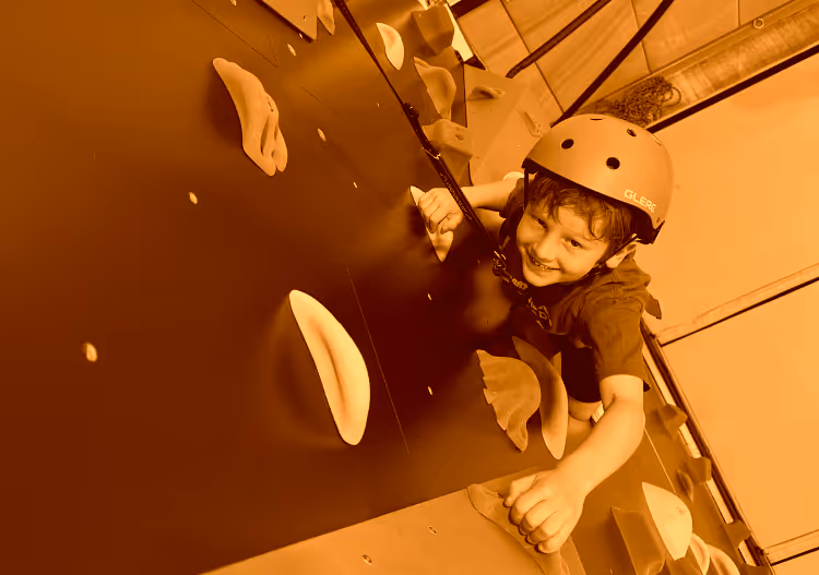 Young boy wearing helmet climbing an indoor rock wall, smiling at the camera.