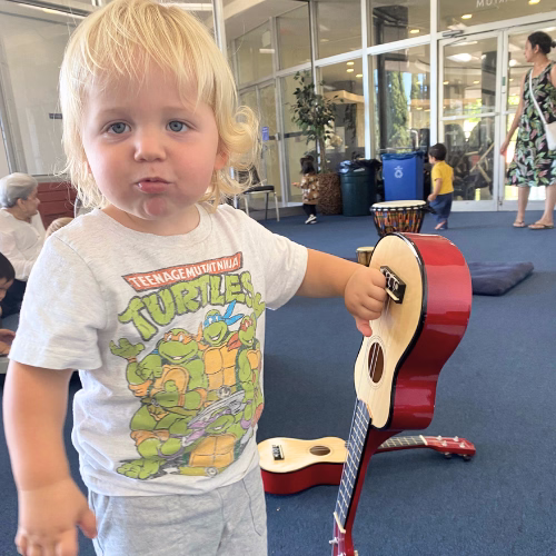 Young blond child wearing a Teenage Mutant Ninja Turtles t-shirt holding a small red guitar in an indoor play area.