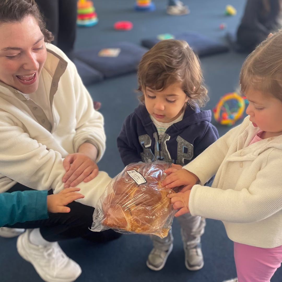Two toddlers touching a plastic bag with bread, accompanied by a smiling adult crouching nearby.