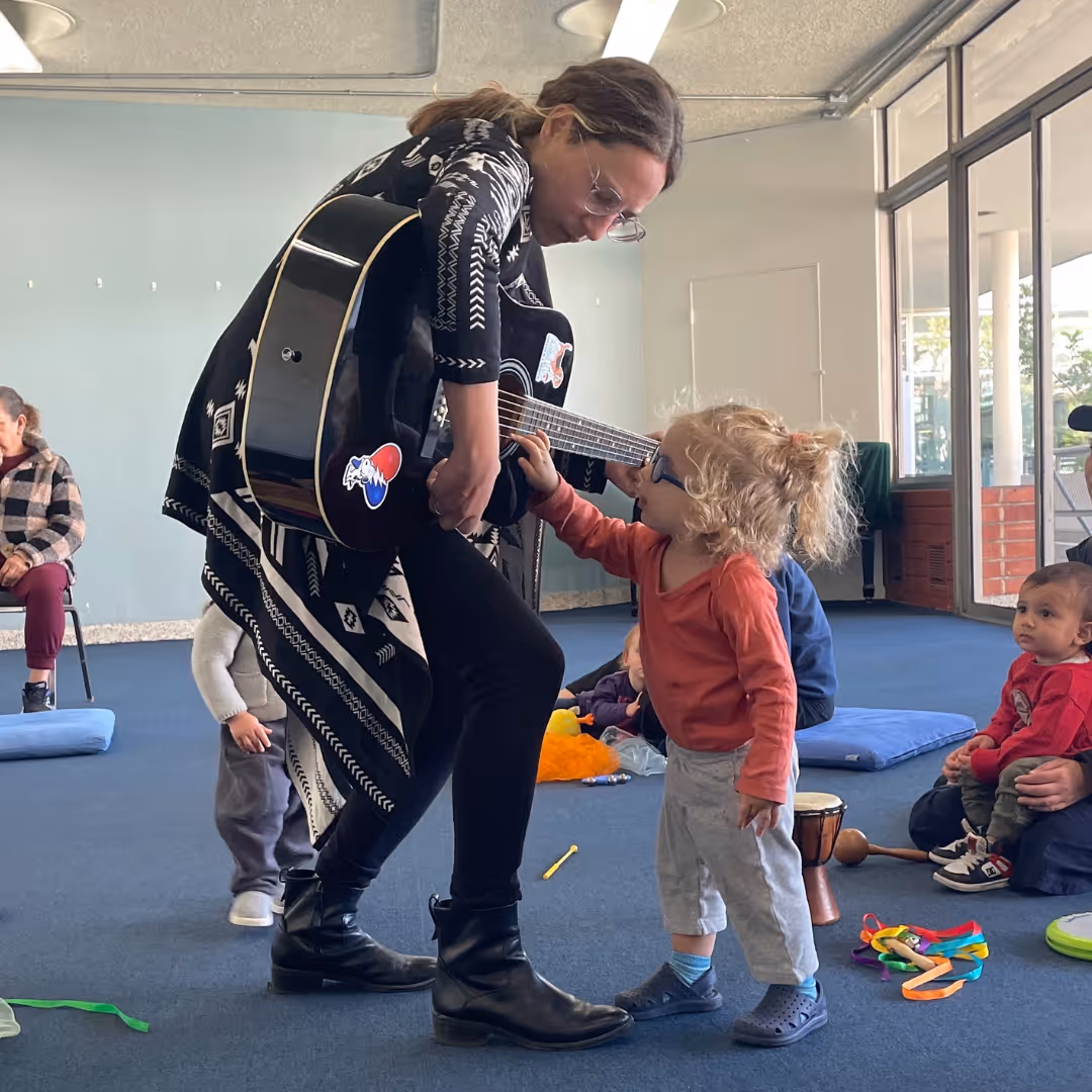 Woman playing guitar and interacting with a young child in a casual indoor setting with other children seated in the background.