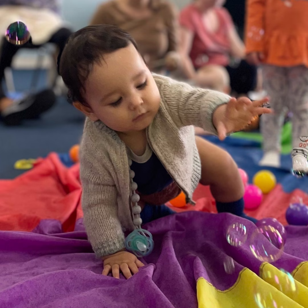 Young child reaching out to catch bubbles while crawling on colorful blankets indoors.