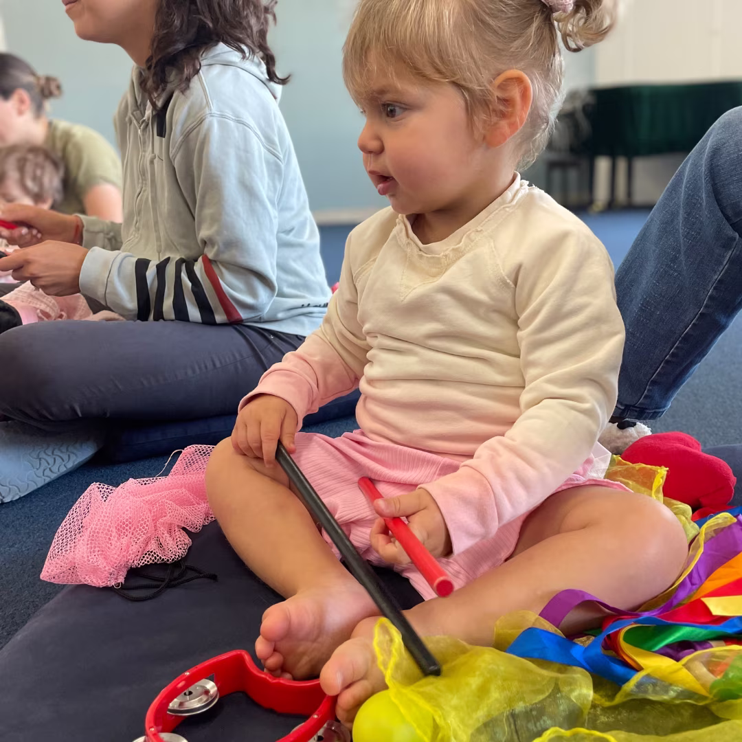 Young child sitting cross-legged on a cushion holding drumsticks and surrounded by colorful scarves and a tambourine.